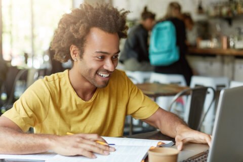 indoor-shot-happy-student-male-with-curly-hair-dressed-casually-sitting-cafeteria-working-with-modern-technologies-while-studying-looking-with-smile-notebook-receiving-message-from-friend