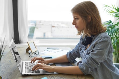 people-modern-technology-job-concept-side-view-serious-concentrated-young-woman-freelancer-with-headphones-around-her-neck-keyboarding-portable-computer-sitting-by-window-wooden-desk